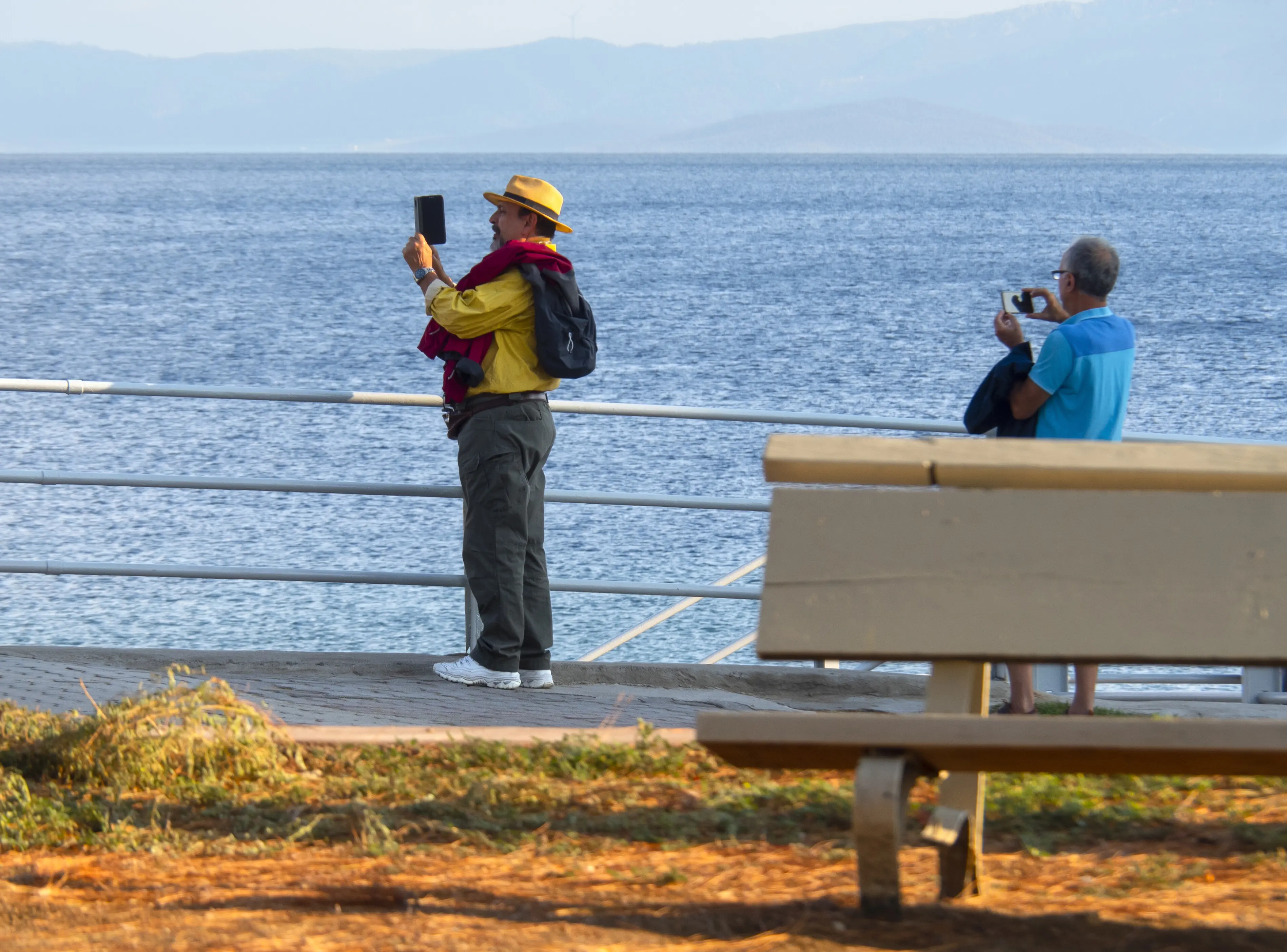 Ein stilvoll gekleideter Mann mit Hut fotografiert auf der Uferpromenade in Griechenland das Meer.